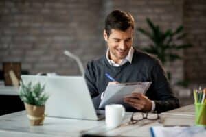 smiling-entrepreneur-writing-business-report-while-working-laptop-office